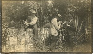 historic photo of men with cannon. 1 man sitting on wall, 2 men looking at cannon. overgrown plants around cannon
