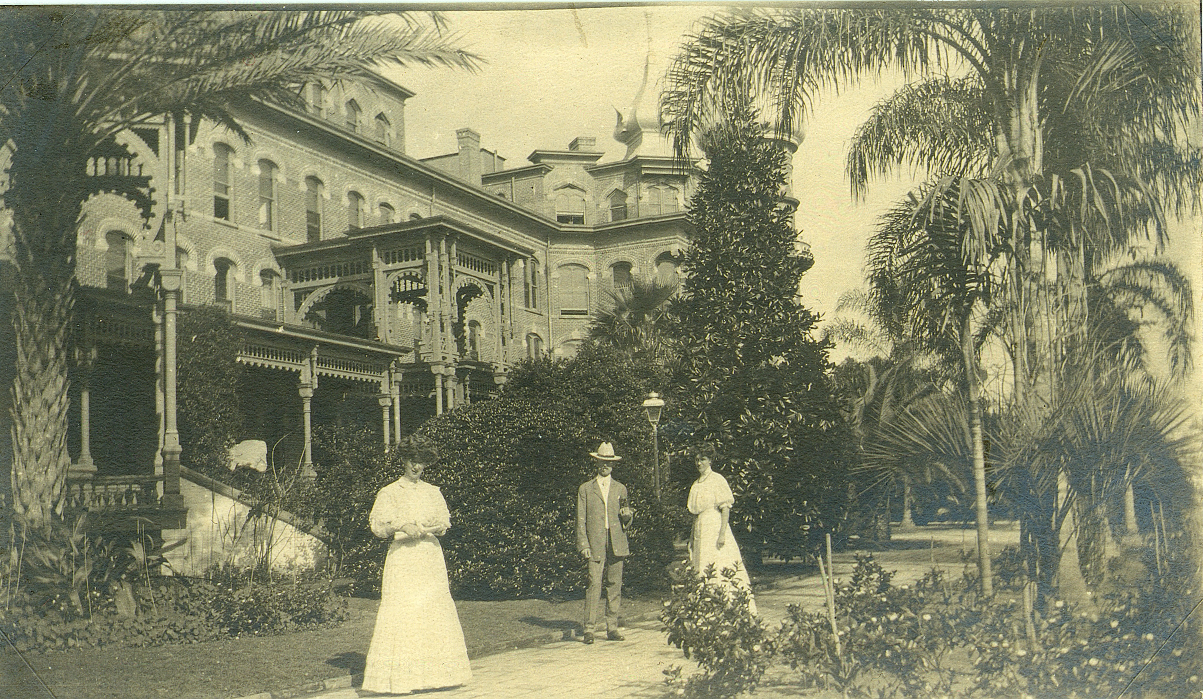 2 women and 1 man in old-fashioned clothing standing in front of hotel, surrounded by lush plants