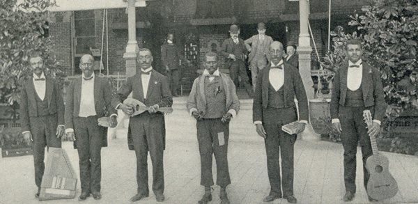 six dark skinned men in front of TBH, posing two feet apart, holding various instruments such as guitar, drum, mandolin. Dressed in minstrel suits with tails, ties and vests.