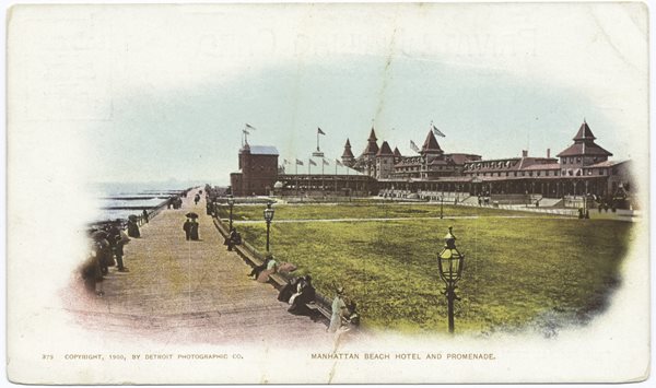 postcard of large building, flags flying, ocean coastline with large boardwalk bordering beach , people strolling and sitting on benches.