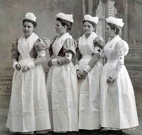 four young women standing dressed in uniform of white apron crossed in front, white folded hats, red cross on armband