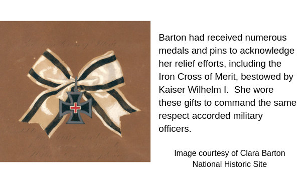 white silk ribbon bow with two black stripes tied with iron cross medallion decorated with small red cross. Caption: Barton had received numerous meals and pins to acknowledge her relief efforts, including the Iron Cross of Merit, bestowed by Kaiser Wilhelm I. She wore these gifts to command the same respect accorded military officers.
