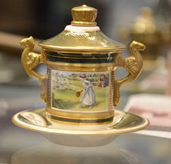 Small porcelain cup and saucer with lid gold trim and glazed image of woman playing tennis .Two opposing handles with winged dragons