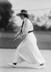 Frances Lippett in long white dress, white shoes dark belt, neck tie and hat swinging a tennis racket