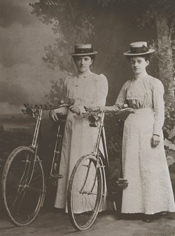 Two women wearing long dresses and hats standing next to bicycles with headlamps