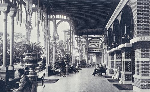 historic photo showing view from one end of East veranda looking down length of veranda. guests lounging in chairs, potted plants on veranda.