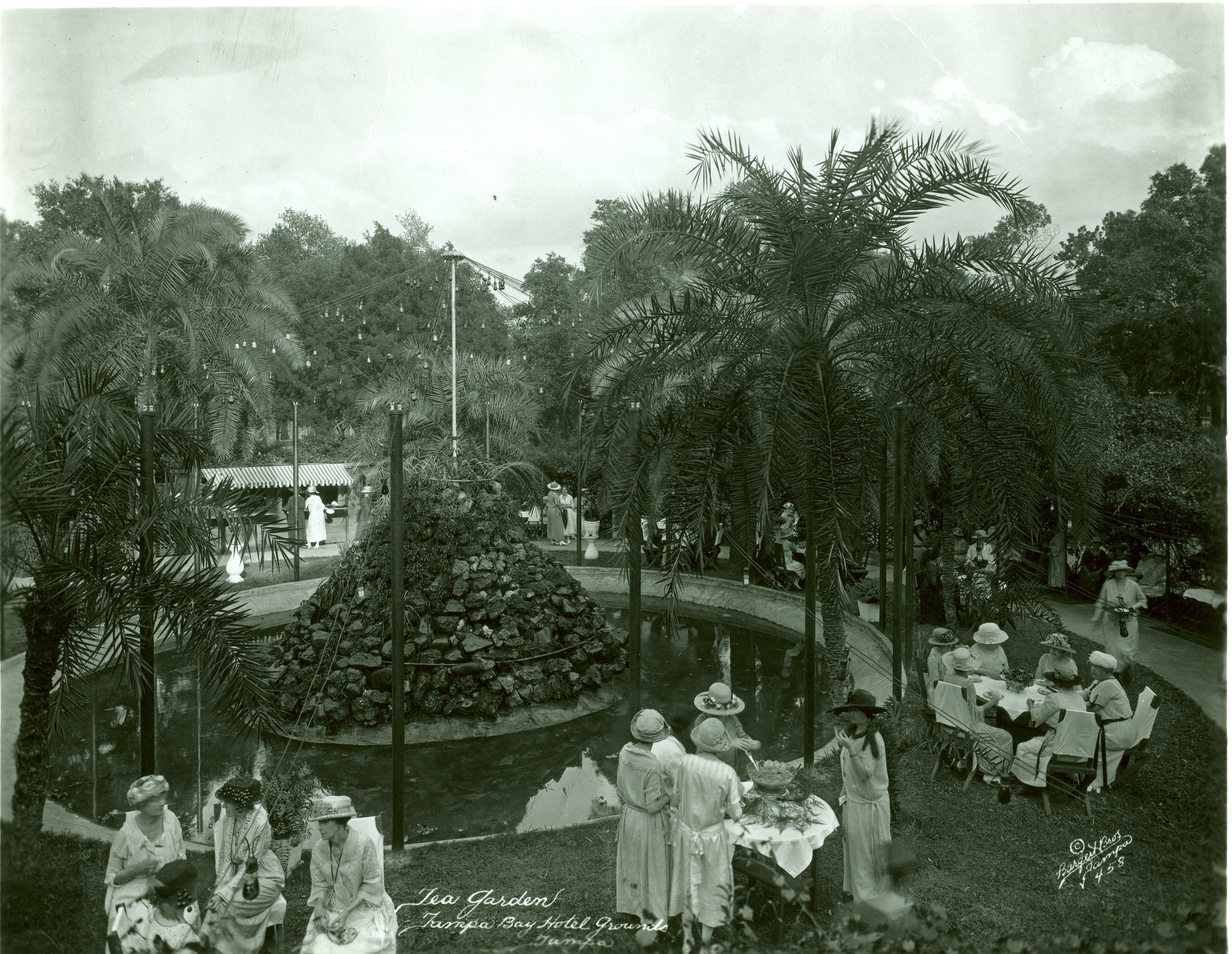 tea garden at Hotel, small round tables with 4-5 ladies at each, fancy dress, large rock fountain in center, lush greenery