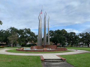 outdoor abstract sculpture of 6 tall metal towers in a circle. American flag flying beyond sculpture