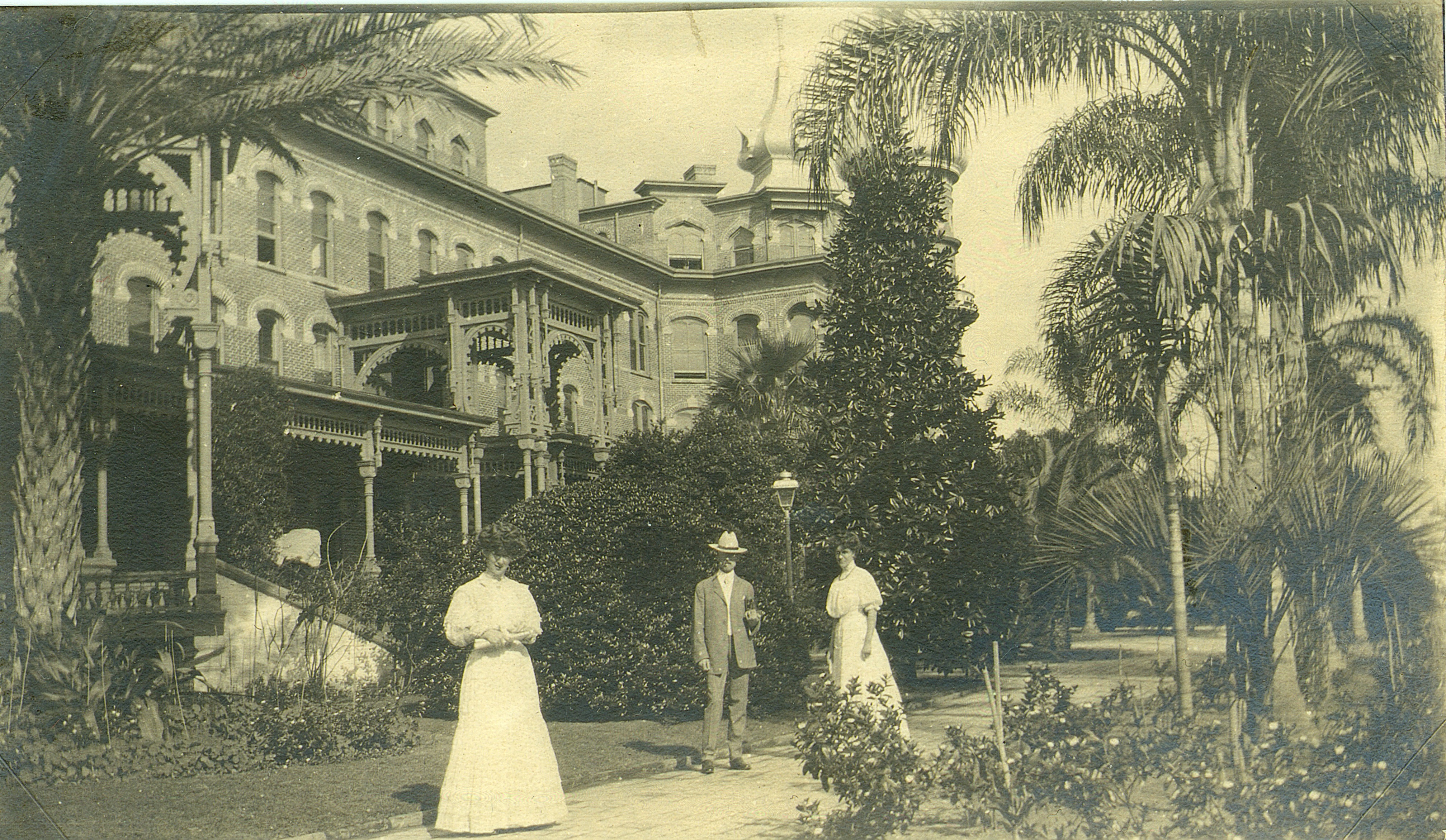 2003-078-015.jpg 2 women and 1 man in old-fashioned clothing standing in front of hotel, surrounded by lush plants