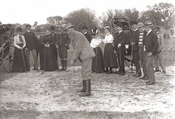 man leaning in suit and high boots leaning forward to swing a golf club, crowd of ladies and gentlemen in semicircle behind watching. Rickshaw parked behind.