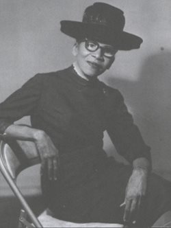 woman seated in chair wearing a dark dress and a medium black hat, looking directly at camera