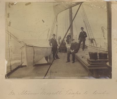 historic photo of men on the deck of a boat, sail above them, lifeboat on the side.