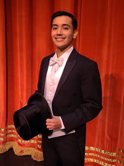 young man in a tuxedo holding a top hat, standing in front of a long red curtain
