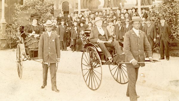Henry and Margaret Plant each in a rickshaw being pulled by dark skinned bellmen. Crowd of people behind viewing from the steps of TBH.