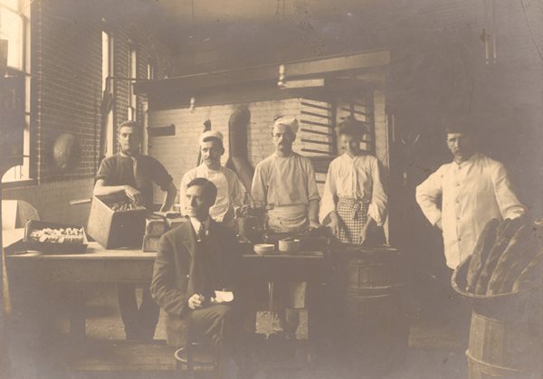 five gentlemen and one lady. Man seated in front dressed in formal suit holding notepad. People behind wearing aprons scarves and hats. Large basket with loaves of bread sitting on a barrel, other baked goods on table.