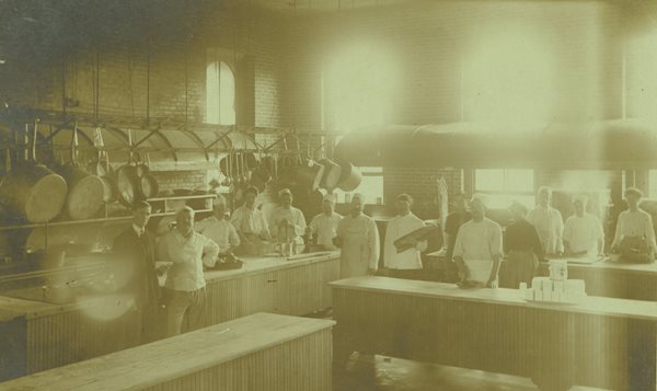 TBH kitchen, chefs and bakers wearing hats and aprons posed in front of large hanging pots, pans, cooking utensils.