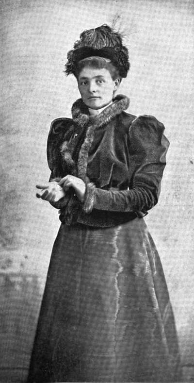 Portrait of Bettina Lesser  standing in a formal dress with  short fur trimmed jacket, mutton sleeves and fur-trimmed collar.