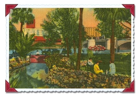creek running through a park full of trees and people in the foreground and a city in the background
