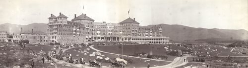 Cars in procession in front of large grand hotel, open field rolling hills and mountain range in background