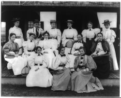 Victorian ladies wearing long dresses with mutton sleeves, holding tennis rackets seated and standing on stair steps