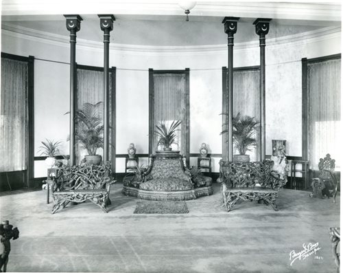 historic photo showing Hotel solarium. two briar root benches set next to ornate pouf. potted palm trees in background.