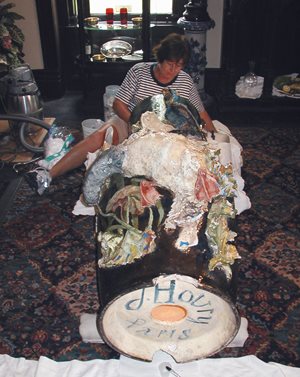 large vase on its side, bottom in foreground. a woman sits on the floor near the top of the vase and performs repairs.