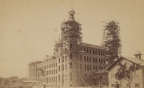 historic photo of hotel under construction. scaffolding surrounds 2 minarets being built, wooden building next to Hotel.