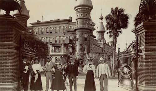 historic photo of 7 Hotel guests standing at gate to Hotel property. 3 ladies wearing long skirts and blouses, men in suits, all wearing hats.