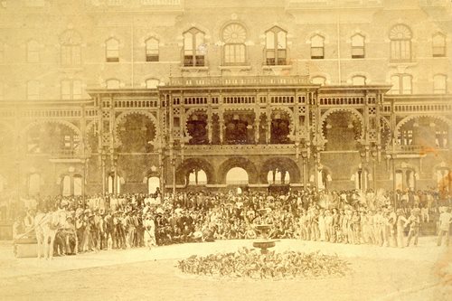 historic photo showing hotel workers outside East veranda. hundreds of men in work clothes and 2 horses visible.