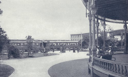 historic photo showing train outside Hotel, veranda in foreground, guests walking to and from train