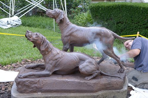 Dog statue, man leaning down near hind legs, blasting legs with dry ice creating steam. Statue is roped off with caution tape.