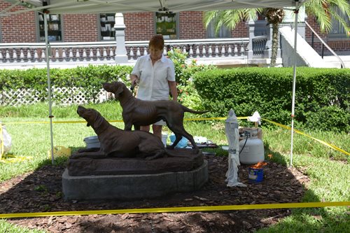 Dog statue, woman standing behind spreading hot wax on statue, supplies and wax tank off to the side. Tent over statue and woman and caution tape roping off area.