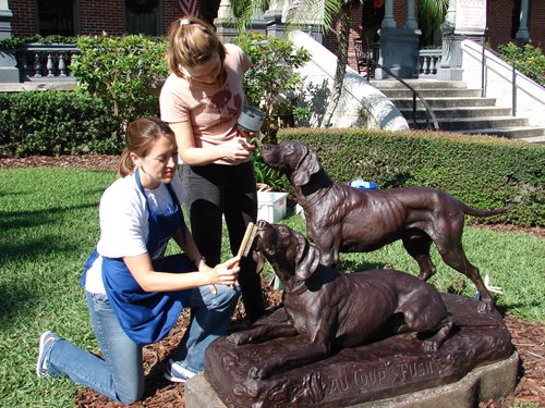 Two women applying cold wax to dog sculpture using brushes and other small tools.