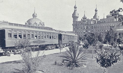 historic photo of train arriving at Hotel. building visible in background with trees and plants along train tracks.